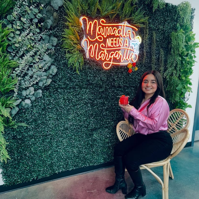 A woman in a pink shirt holds a red drink, sitting by a neon "Mamacita Needs a Margarita" sign.
