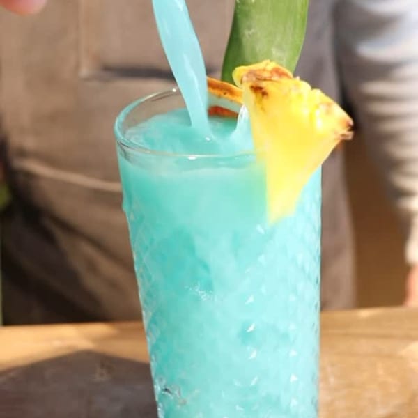 Blue margarita being poured into a tall glass, garnished with a pineapple slice and green leaf.