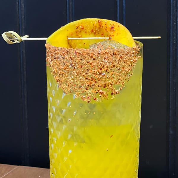 Close-up of a yellow margarita with a chili-lime rim and a mango slice garnish.