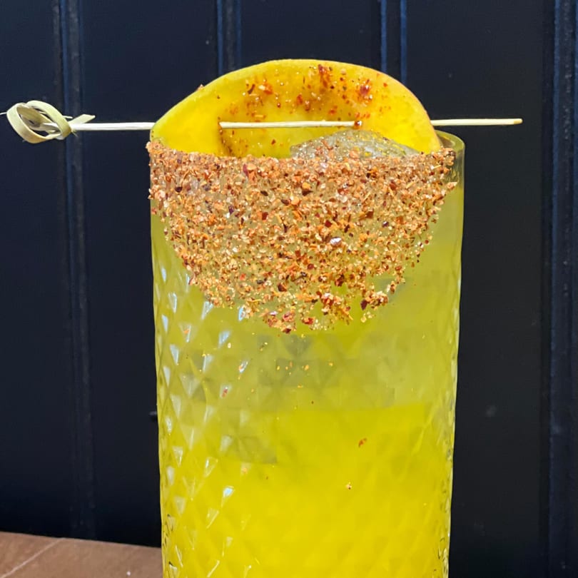 Close-up of a yellow margarita with a chili-lime rim and a mango slice garnish.