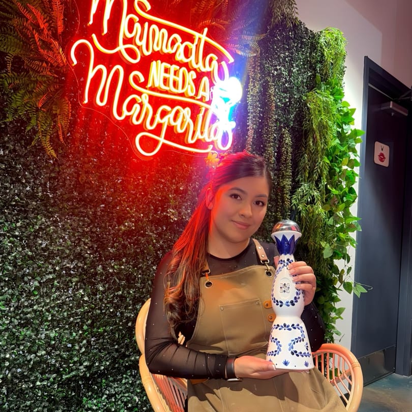 A woman in an apron holds a decorative tequila bottle in front of a neon sign and plant wall.