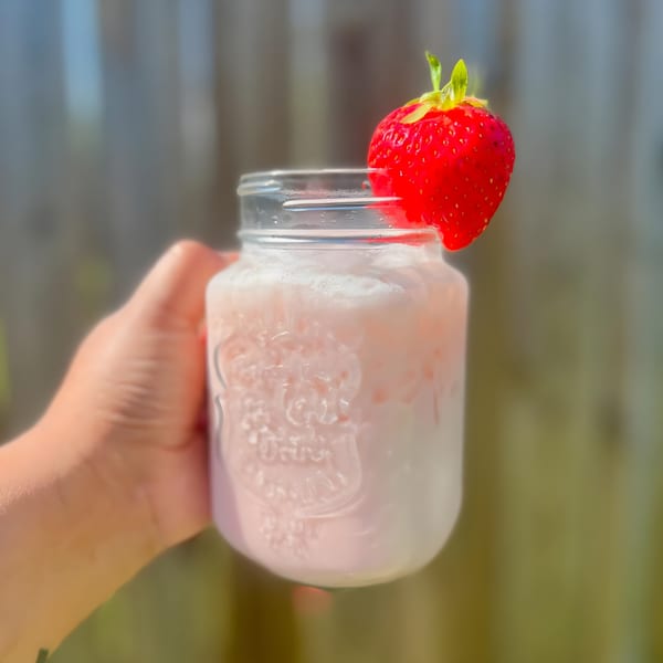 A hand holds a mason jar filled with a pink drink, garnished with a fresh strawberry.