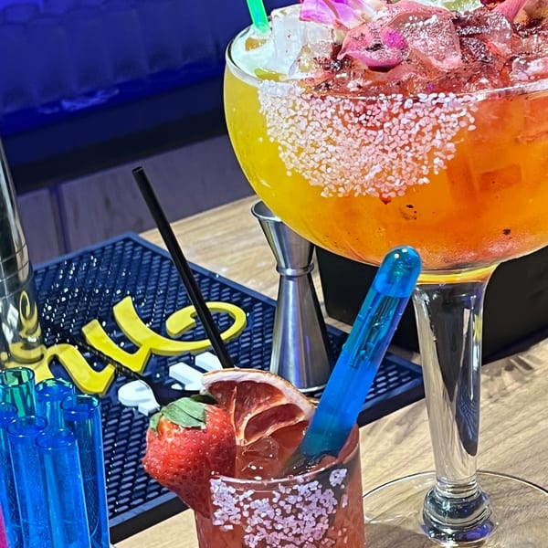 Two vibrant margaritas, one large orange and one smaller red, garnished with fruit and salt.