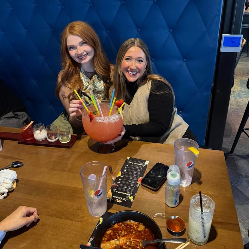 Two smiling women at Los Compas Margaritatown sharing a giant strawberry margarita with straws.