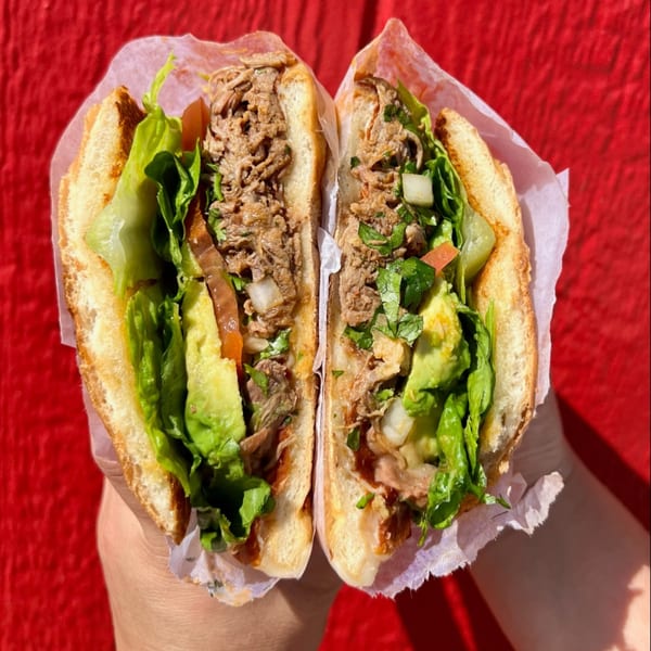 A Mexican torta sandwich, cut in half, filled with meat, lettuce, avocado, and cilantro.