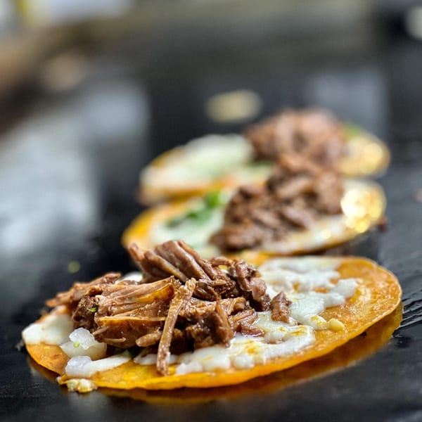 Three Los Compas Margaritatown tacos with shredded meat and white cheese on a dark surface.