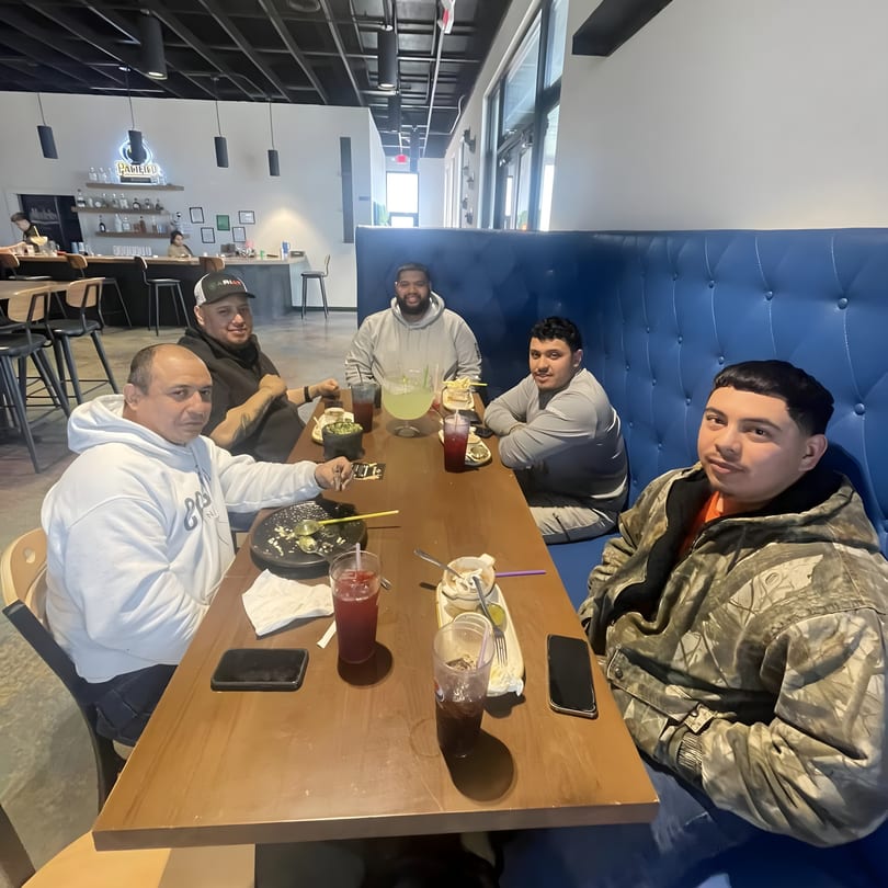 Five men seated at a long table with drinks and food, enjoying a meal at Los Compas Margaritatown.