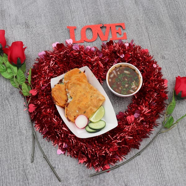 Heart-shaped birria tacos with consommé, radish, cucumber, and lime, surrounded by red tinsel and roses.