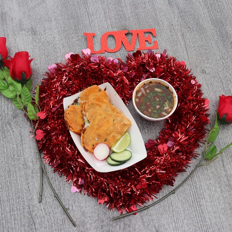 Heart-shaped birria tacos with consommé, radish, cucumber, and lime, surrounded by red tinsel and roses.