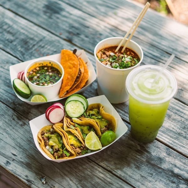 Mexican food spread on a rustic wooden table, featuring tacos, birria, and a green drink.