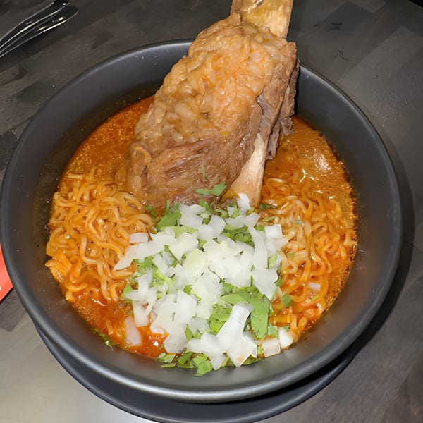 A large bone-in meat dish with noodles, broth, diced onions, and cilantro in a dark bowl.